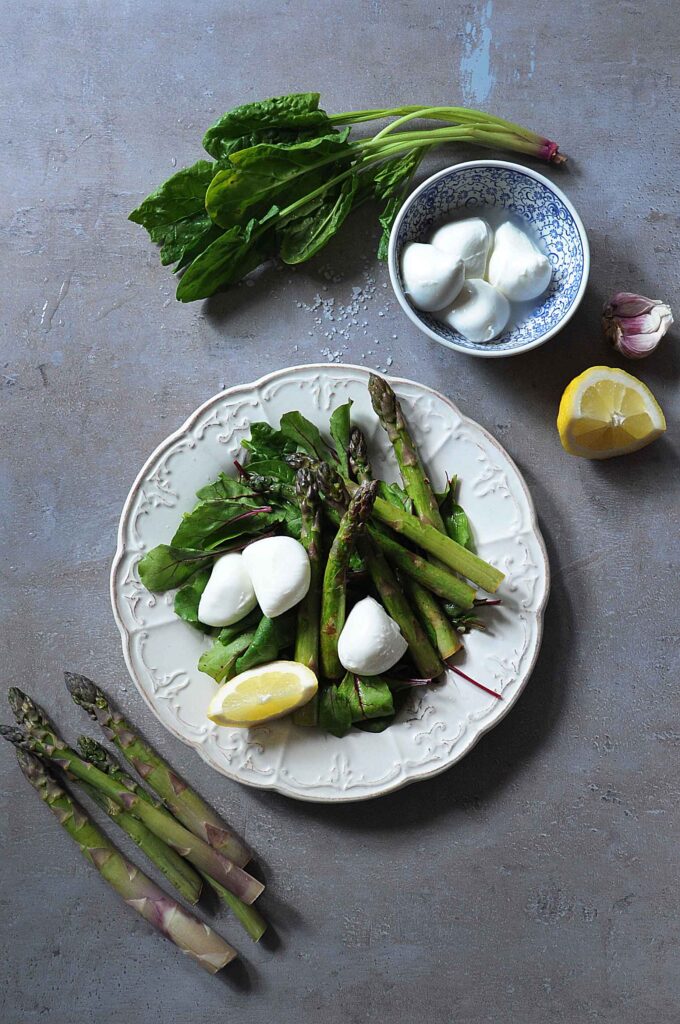 Overhead view of a warm asparagus salad with fresh mozzarella, leafy greens, and lemon on a serving plate.