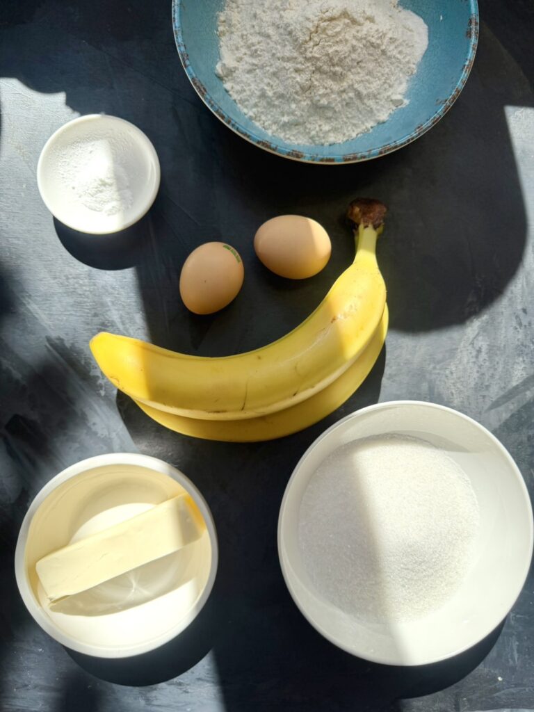 Ingredients for homemade banana bread including ripe bananas, flour, eggs, butter, sugar, baking powder, and baking soda.
