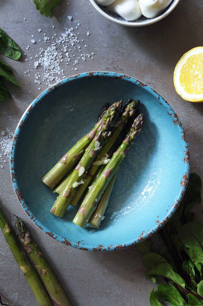 Cooked asparagus spears on a blue plate with sea salt and lemon for a warm asparagus salad.