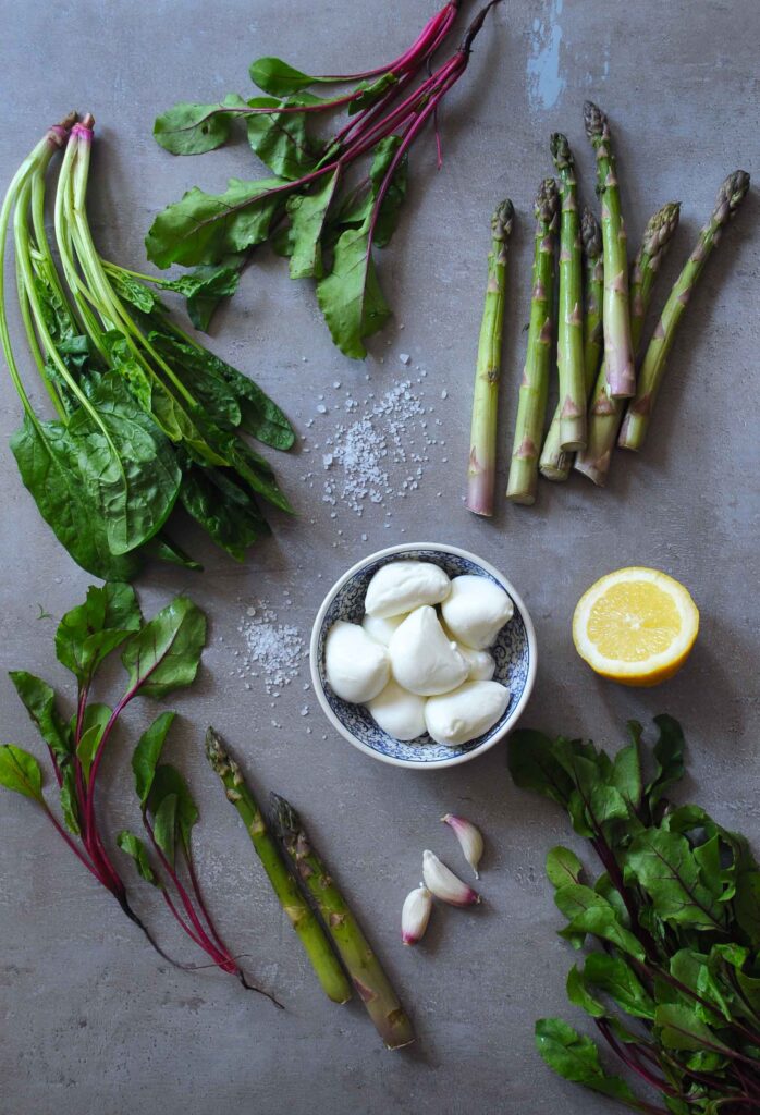 Fresh ingredients for warm asparagus salad with mozzarella, lemon, garlic, and leafy greens.