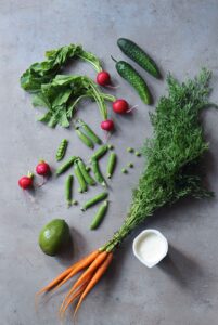 Fresh ingredients for avocado Greek yogurt dip including avocado, carrots, cucumbers, radishes, snap peas, and Greek yogurt.