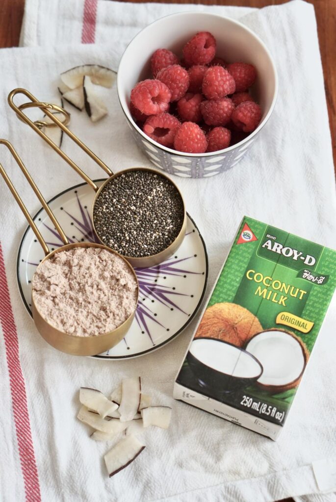 Flat lay of protein chia pudding ingredients including chia seeds, protein powder, coconut milk, coconut flakes, and fresh raspberries