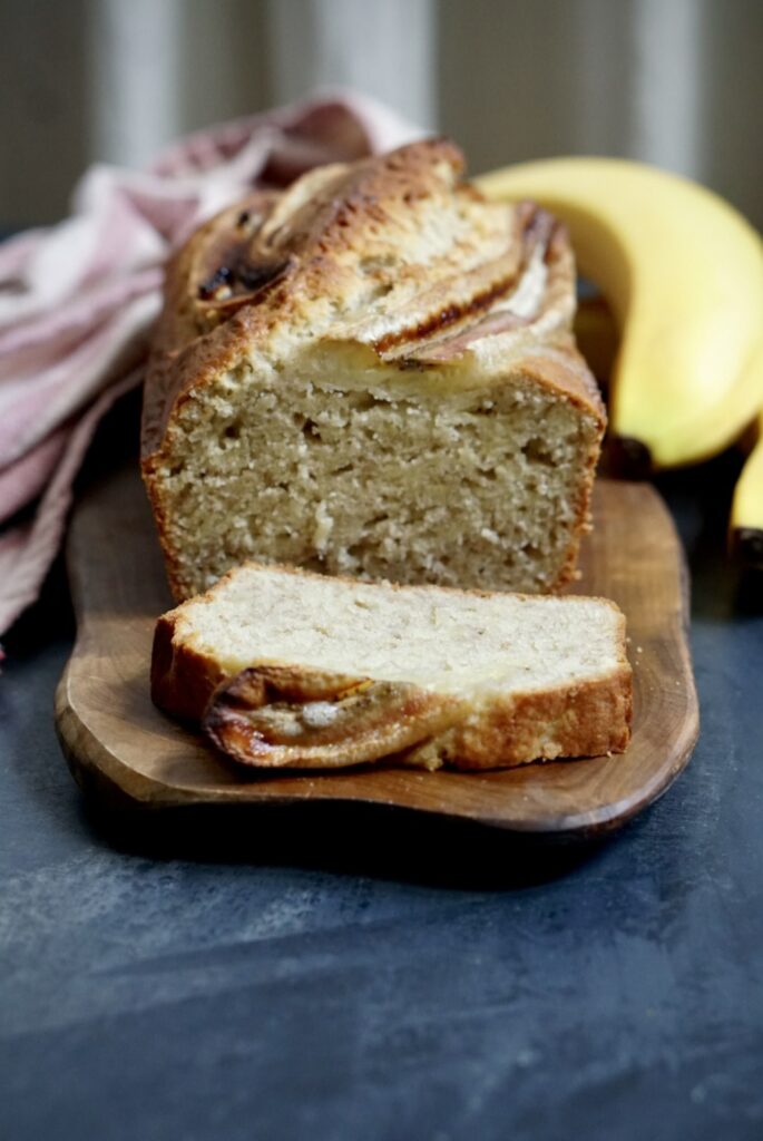 Freshly baked homemade banana bread sliced on a wooden board with ripe bananas in the background.