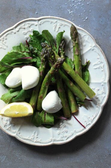Warm asparagus salad with fresh mozzarella, leafy greens, and lemon served on a white plate.