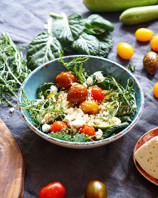 Bowl of warm grilled zucchini and cherry tomato salad topped with sheep cheese and fresh greens