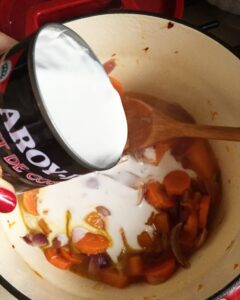 Pouring coconut milk into a pot of cooked pumpkin, carrot, and onions