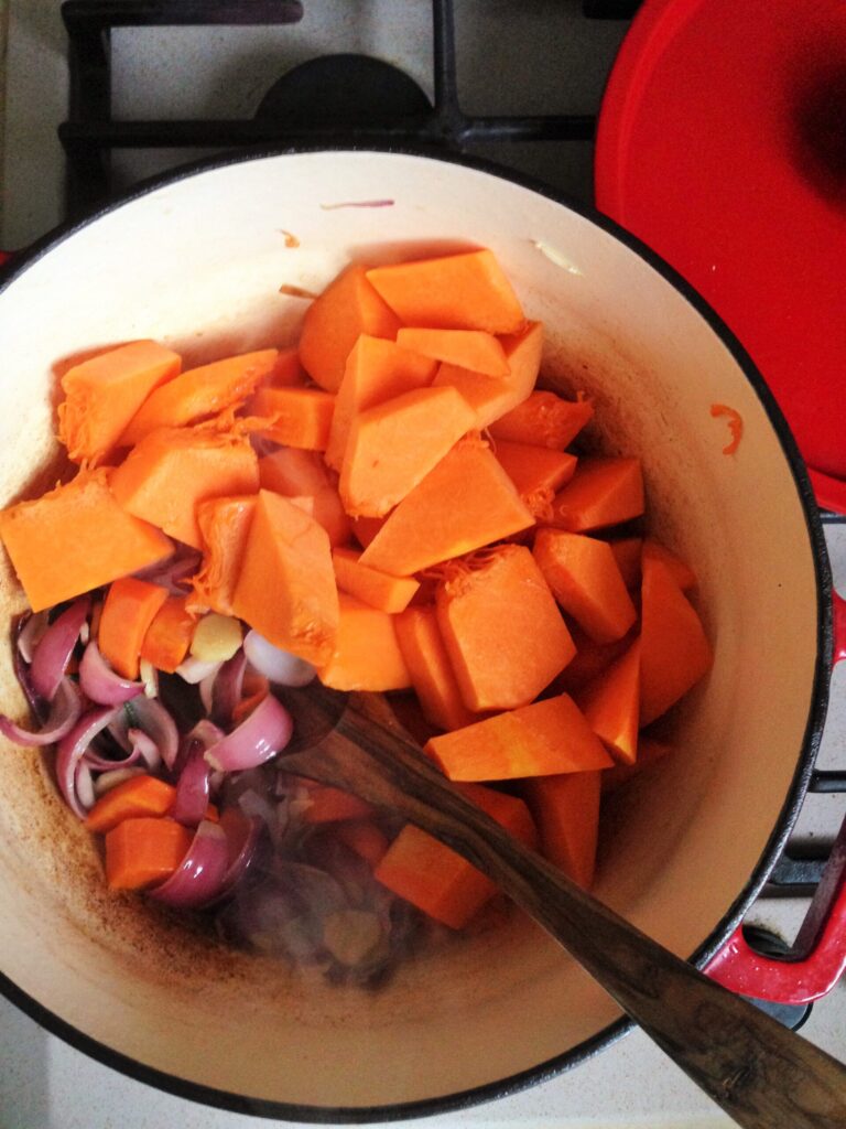 A top-down view of a red pot on a stovetop filled with chunks of raw orange pumpkin and sliced red onions being sautéed with a wooden spoon.