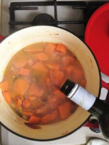 A close-up of a dark green bottle pouring white wine into a red pot filled with simmering chunks of pumpkin, carrots, and broth.