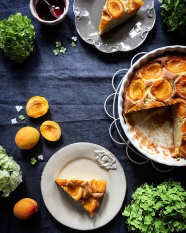 Overhead view of a partially sliced apricot cake in a white fluted baking dish, with two slices served on rustic plates, fresh apricots, a small bowl of jam, and green hydrangeas on a dark blue linen cloth.
