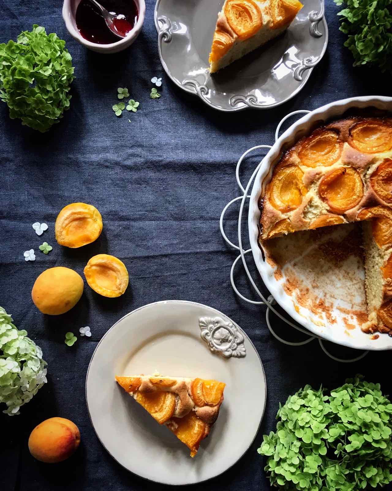 Overhead view of a partially sliced apricot cake in a white fluted baking dish, with two slices served on rustic plates, fresh apricots, a small bowl of jam, and green hydrangeas on a dark blue linen cloth.