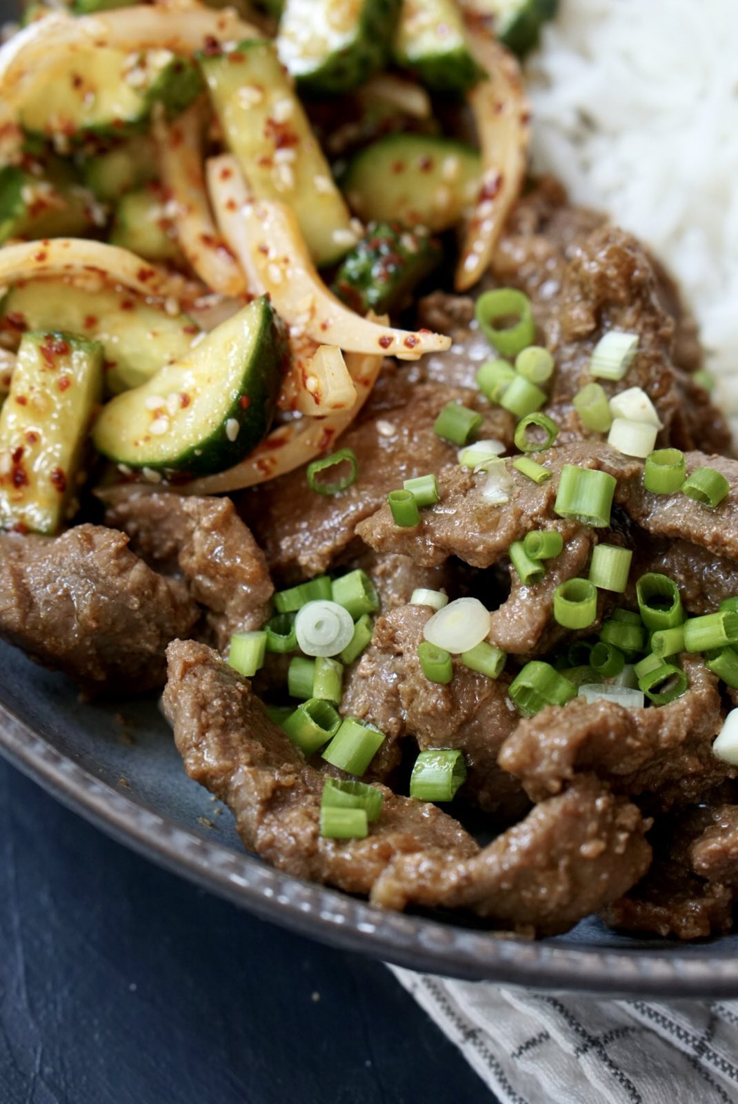Close-up of beef stir-fry with soy sauce, green onions, white rice, and spicy cucumber salad.