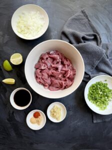 Overhead view of raw sliced beef, chopped onion, garlic, green onions, soy sauce, and spices arranged for stir-fry