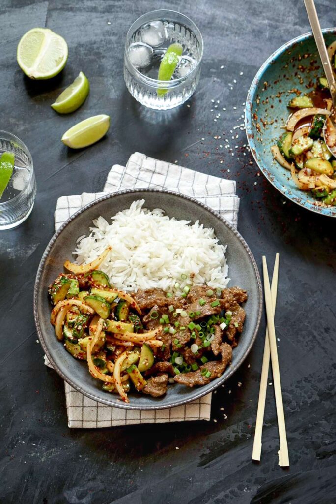 Plate of beef stir-fry topped with green onions, served with rice, chopsticks, and lime wedges on the side