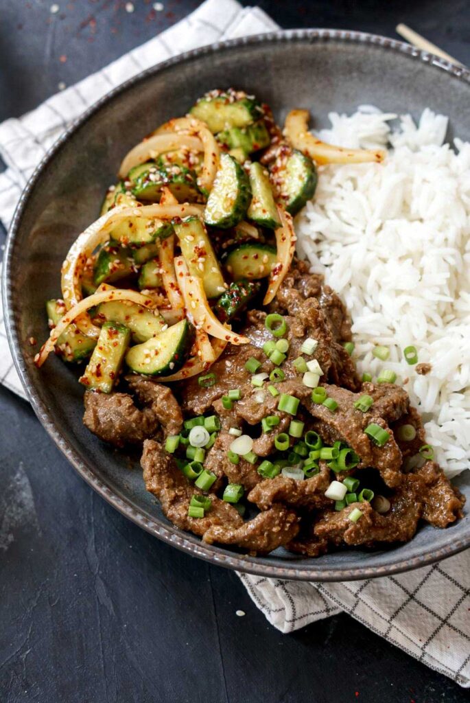 Bowl of Asian-style beef stir-fry served with rice and a cucumber onion salad, garnished with green onions