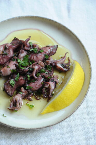 Cooked baby squid garnished with fresh parsley and a large lemon wedge on a white plate
