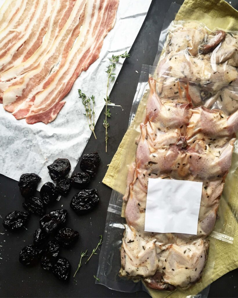 Final presentation of a baked bacon-wrapped quail stuffed with prunes. The crispy, golden-brown quail is served on an elegant vintage-style grey plate, accompanied by fresh cherry tomato halves and sprouts, resting on a yellow napkin over a dark background.