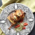 A close-up of a plated, oven-baked quail wrapped in crispy bacon and stuffed with prunes, served on an ornate grey plate with cherry tomatoes and microgreens.