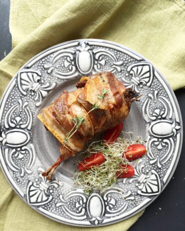 A close-up of a plated, oven-baked quail wrapped in crispy bacon and stuffed with prunes, served on an ornate grey plate with cherry tomatoes and microgreens.