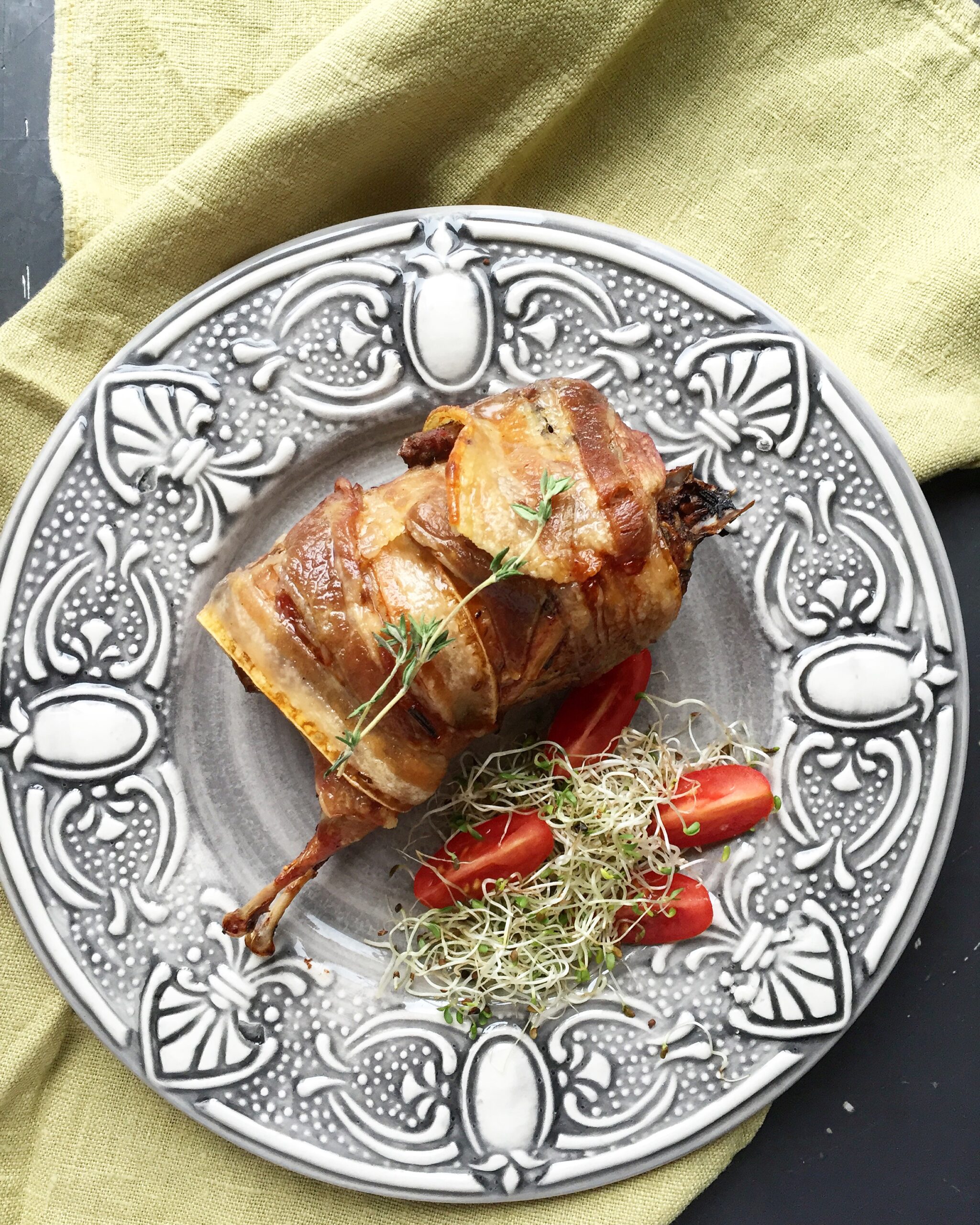 A close-up of a plated, oven-baked quail wrapped in crispy bacon and stuffed with prunes, served on an ornate grey plate with cherry tomatoes and microgreens.