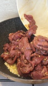 Marinated beef strips being poured into a hot skillet with garlic, ready to sear