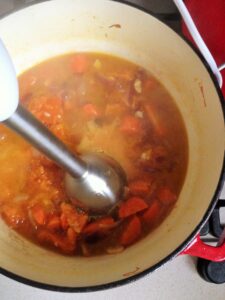 A close-up looking into a red pot where an immersion blender is resting in a chunky, bright orange pumpkin soup mixture with cooked carrots and onions, ready to be pureed.