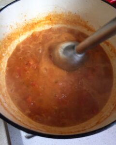 Using an immersion blender to puree cooked tomatoes and red bell peppers in a pot for a creamy soup.