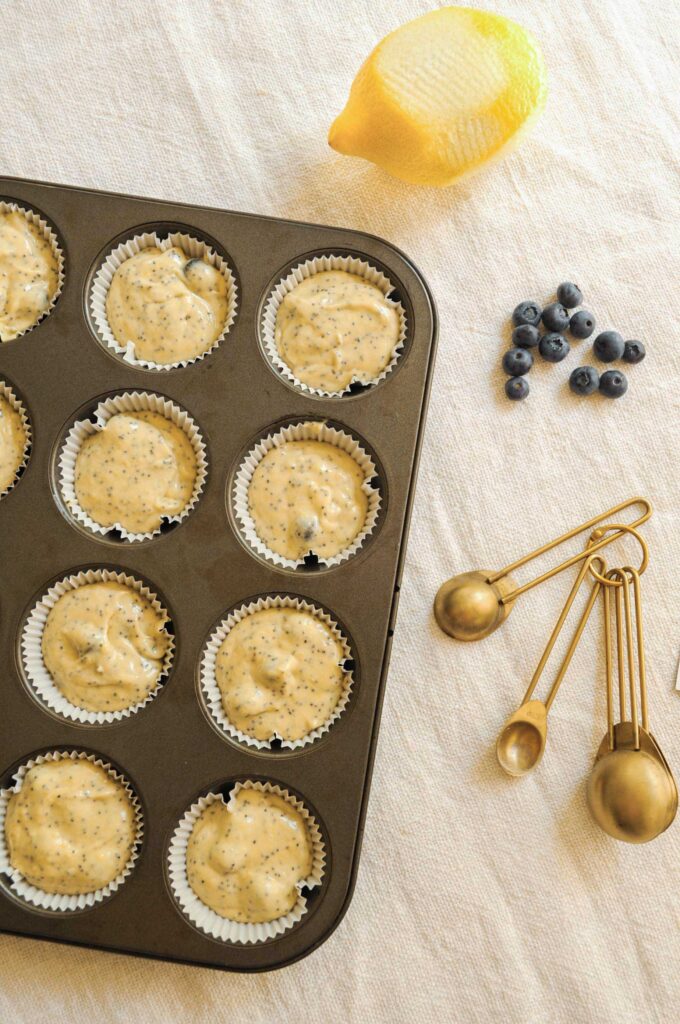 Muffin batter with poppy seeds in a lined muffin pan, next to a fresh lemon, blueberries, and gold measuring spoons