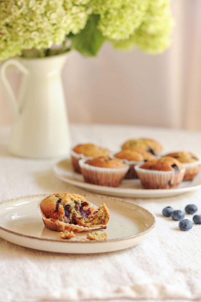 A plate of blueberry lemon poppy seed muffins with one muffin broken open, showing the soft crumb inside, styled with fresh blueberries and flowers