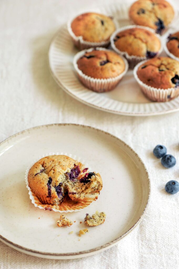 Blueberry lemon poppy seed muffins on a plate, with one muffin broken open to show the moist crumb studded with blueberries and poppy seeds