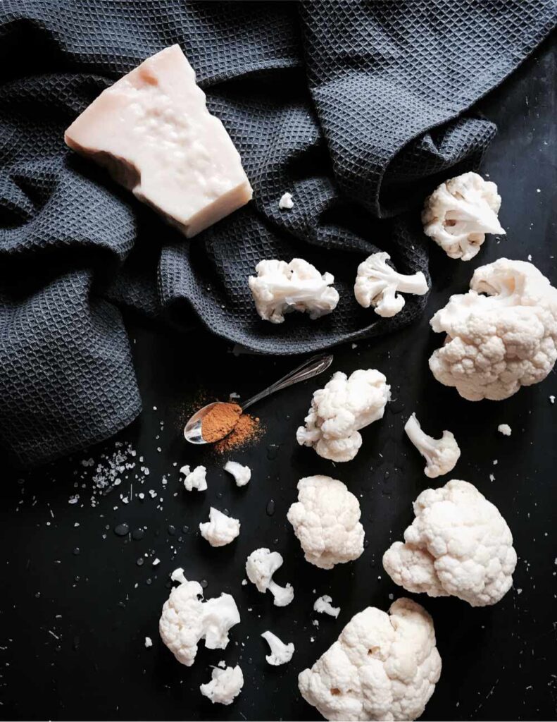 Creamy cauliflower soup with Parmesan ingredients: raw cauliflower florets, a block of Parmesan cheese, and ground spices on a dark surface.
