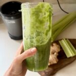 A hand holding a tall glass of celery apple green smoothie with ginger, with fresh celery and ginger visible on the counter behind it