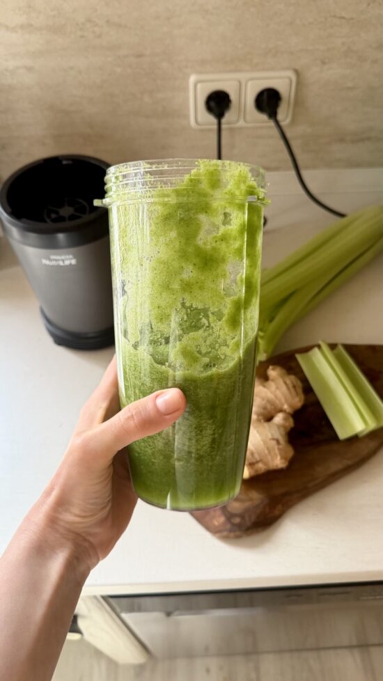 A hand holding a tall glass of celery apple green smoothie with ginger, with fresh celery and ginger visible on the counter behind it