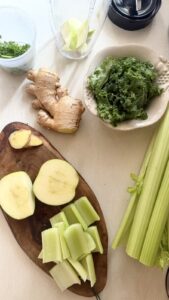 Sliced green apple, celery, ginger, and kale on a cutting board next to a blender, prepped for making a celery apple green smoothie with ginger