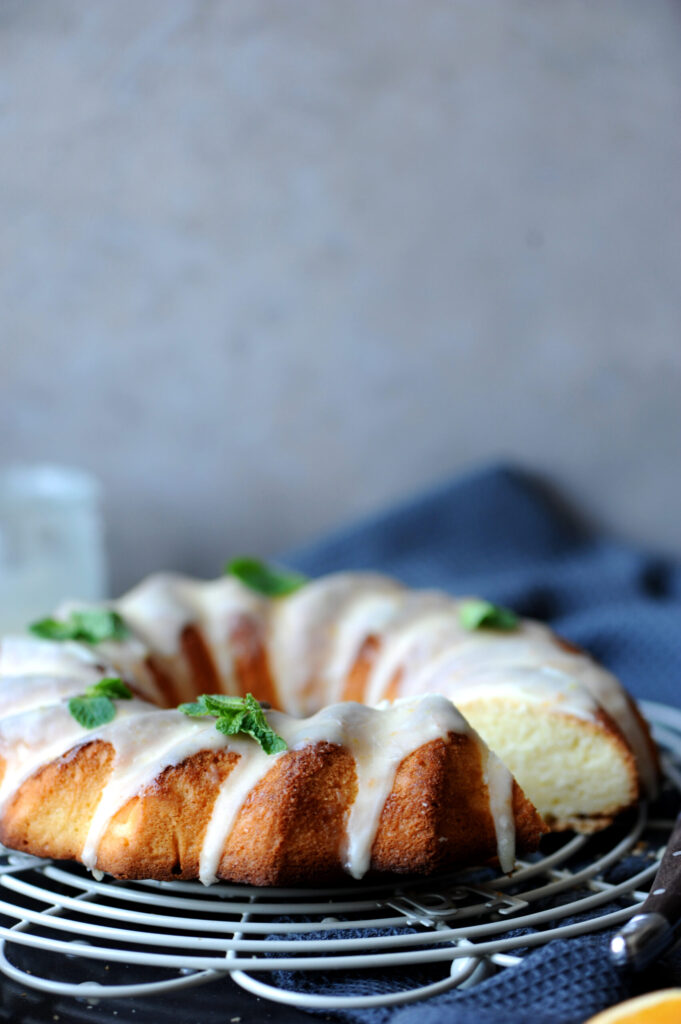 Whole citrus lemon ginger bundt cake with white orange glaze dripping down the sides, garnished with fresh mint leaves on a wire cooling rack.