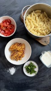 Pot of cooked fusilli pasta al dente next to cooked chicken, tomatoes, herbs, and cream