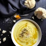 A top-down view of a white bowl filled with creamy cauliflower soup, garnished with a drizzle of olive oil, herbs, and shaved Parmesan cheese. It sits on a dark background next to a blue linen napkin and fresh raw cauliflower florets.