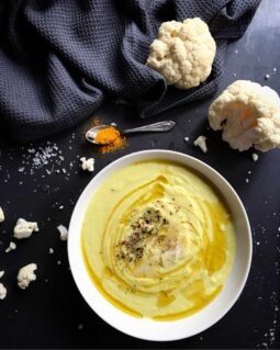 A top-down view of a white bowl filled with creamy cauliflower soup, garnished with a drizzle of olive oil, herbs, and shaved Parmesan cheese. It sits on a dark background next to a blue linen napkin and fresh raw cauliflower florets.