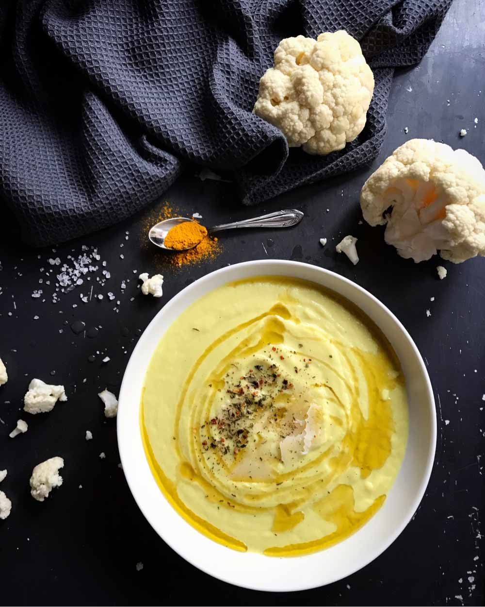 A top-down view of a white bowl filled with creamy cauliflower soup, garnished with a drizzle of olive oil, herbs, and shaved Parmesan cheese. It sits on a dark background next to a blue linen napkin and fresh raw cauliflower florets.