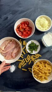 Flat-lay of ingredients for creamy chicken pasta: raw chicken, tomatoes, garlic, cream, herbs, and dry fusilli