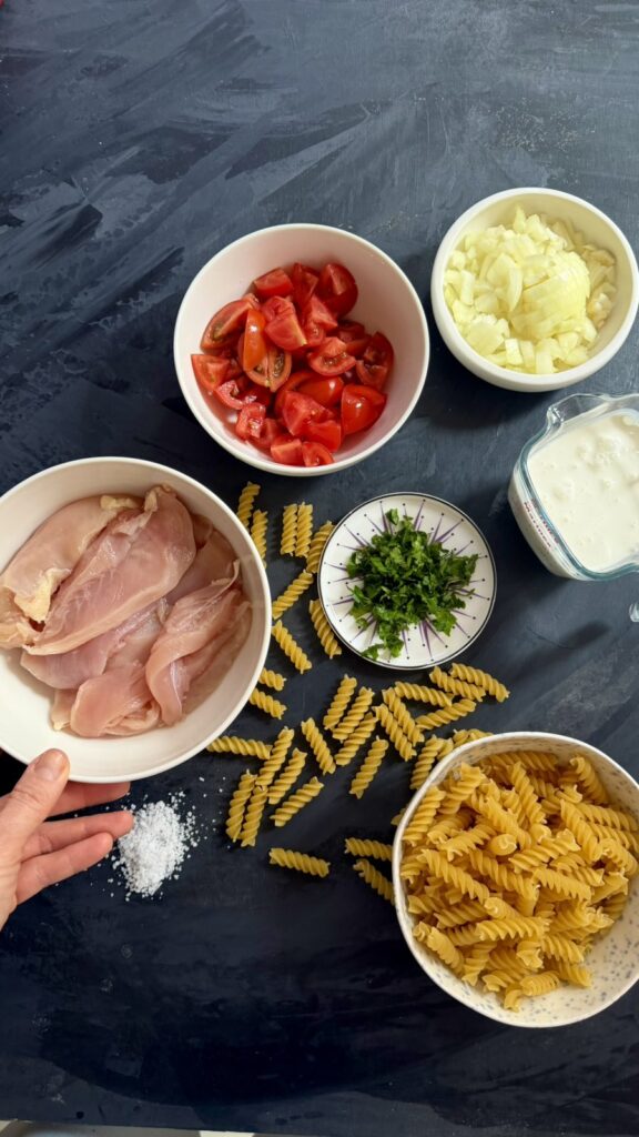 Flat-lay of ingredients for creamy chicken pasta: raw chicken, tomatoes, garlic, cream, herbs, and dry fusilli