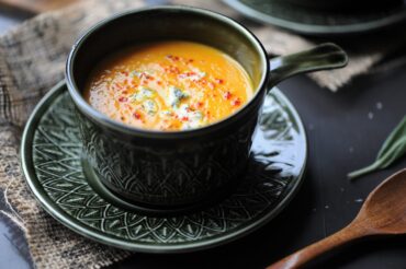 A close-up of a rustic dark green mug filled with creamy pumpkin soup, garnished with crumbled blue cheese and spices, sitting on a matching saucer next to a wooden spoon.