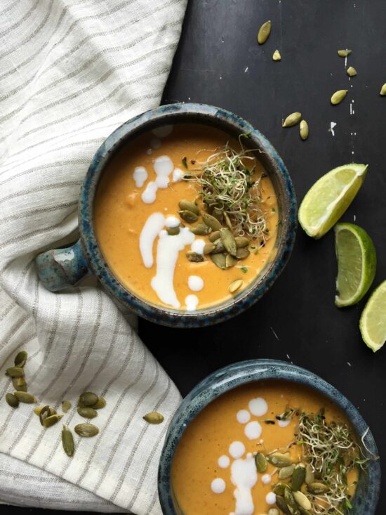 Two bowls of creamy pumpkin soup with coconut milk, topped with pumpkin seeds and alfalfa sprouts