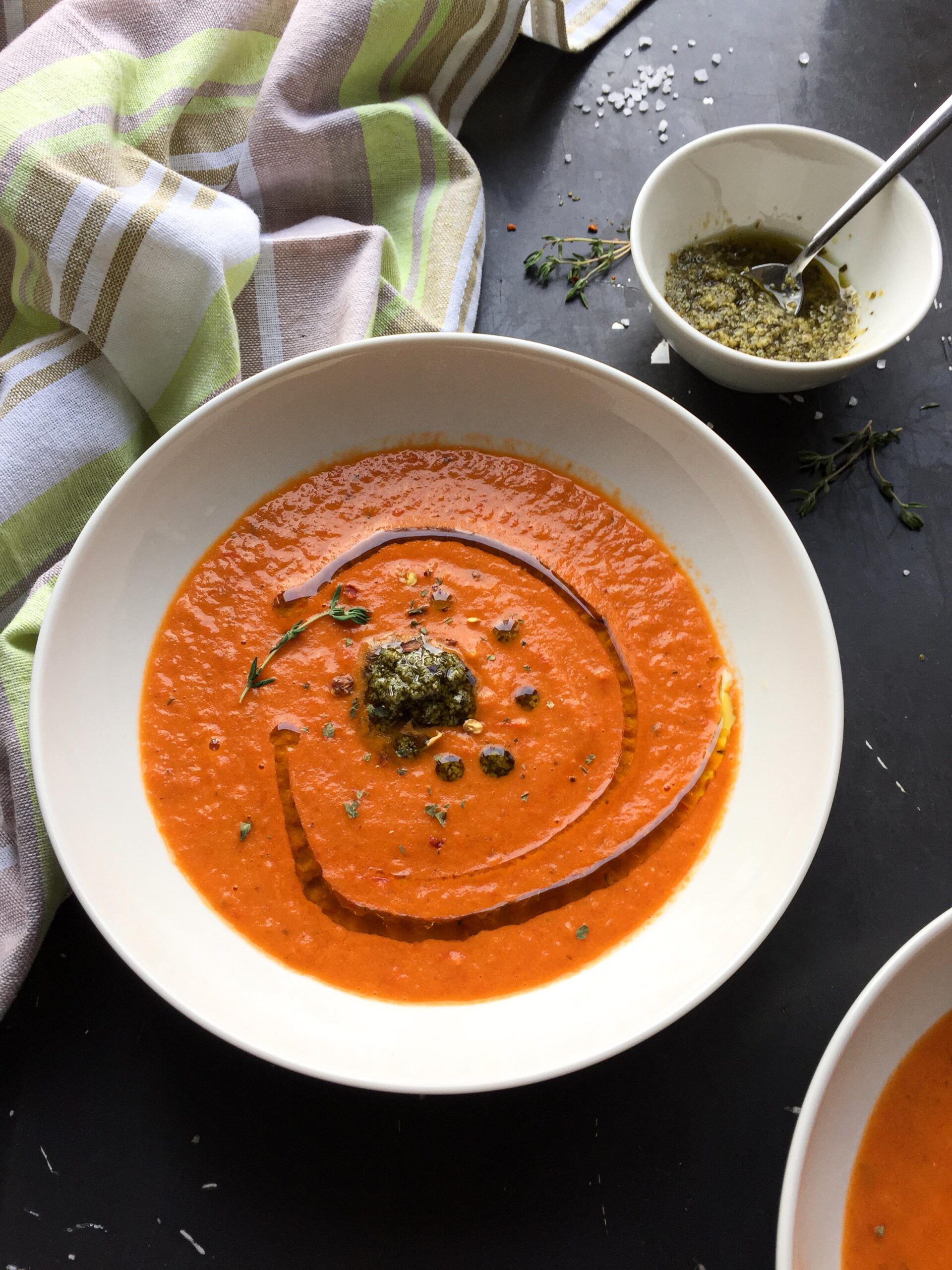 A white bowl of creamy tomato and red pepper soup garnished with a dollop of pesto, fresh thyme, and a dark swirl, sitting on a dark surface next to a striped towel.