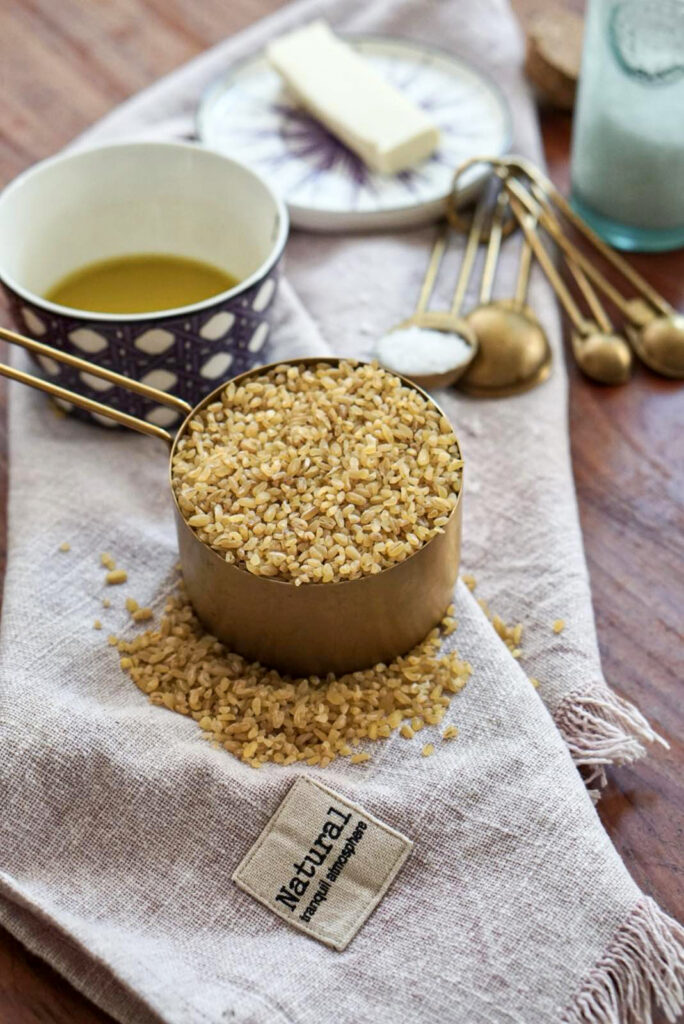 Dry bulgur wheat in a gold measuring cup on a rustic linen napkin, with olive oil and butter in the background.