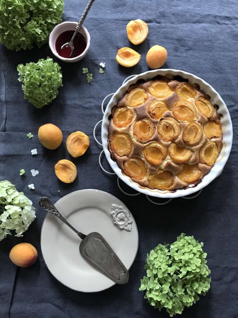 Overhead view of a whole fresh apricot cake in a white fluted baking dish on a dark blue cloth, surrounded by fresh apricots, green hydrangeas, a small bowl of jam, and a white plate with a vintage cake server.