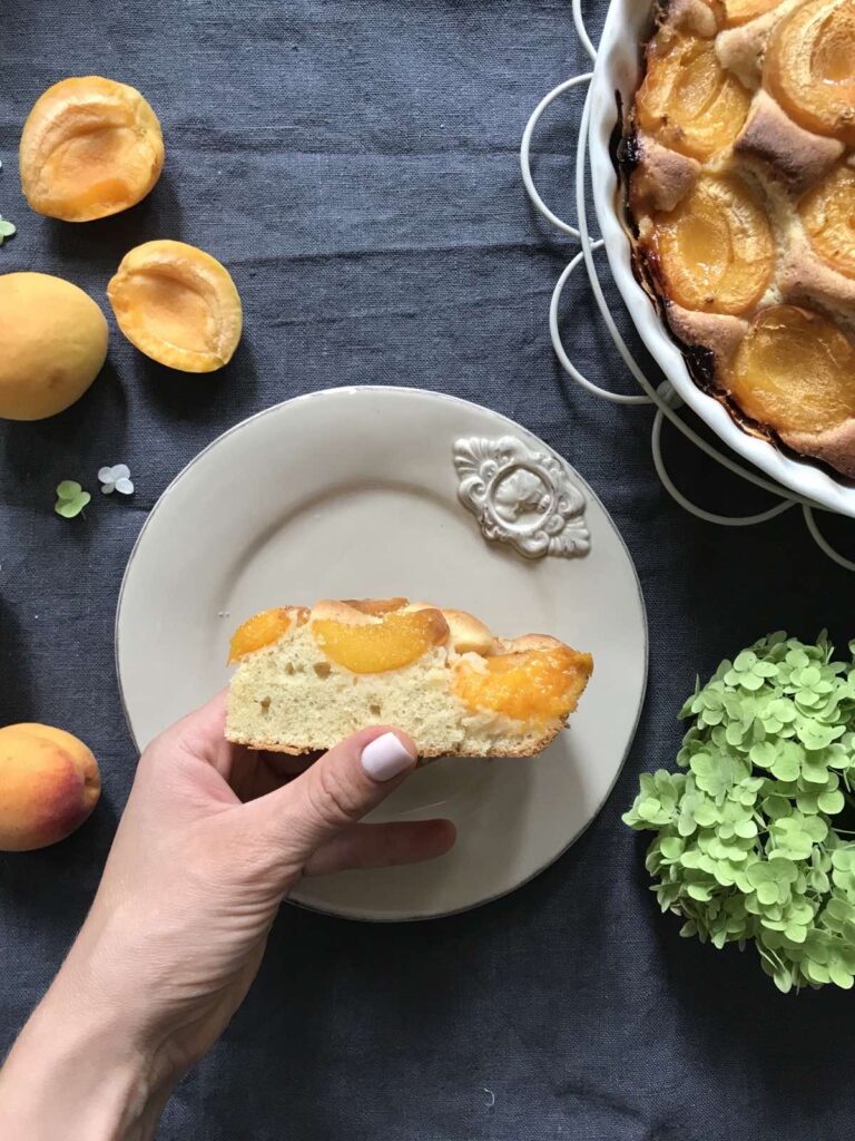 A woman's hand holding a slice of fresh apricot cake over a plate, alongside a white baking dish with the remaining cake, fresh apricots, and a green hydrangea on a dark blue cloth.