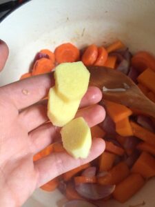 Hand holding peeled fresh ginger over a pot of carrots and onions for pumpkin soup