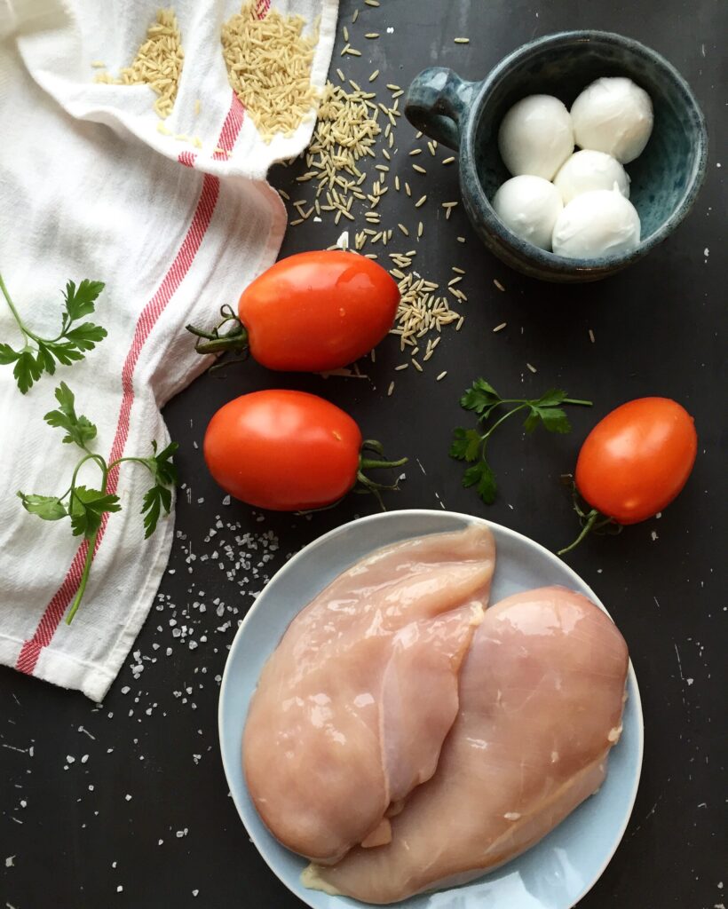 Fresh slices of mozzarella cheese, ripe red tomatoes, and raw chicken breasts on a prep board