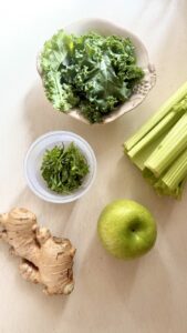 Fresh celery, green apple, ginger root, and kale arranged on a counter, the key ingredients for a celery apple green smoothie with ginger