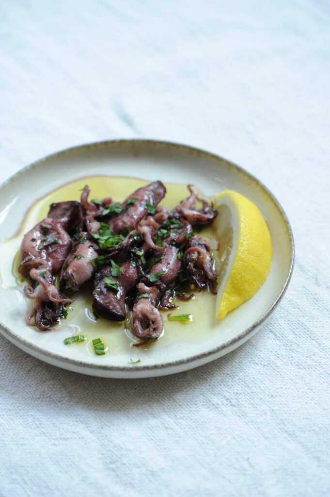 Detailed view of a plate of grilled baby squid garnished with chopped fresh parsley and olive oil, served with a large fresh lemon wedge on a light blue linen surface.